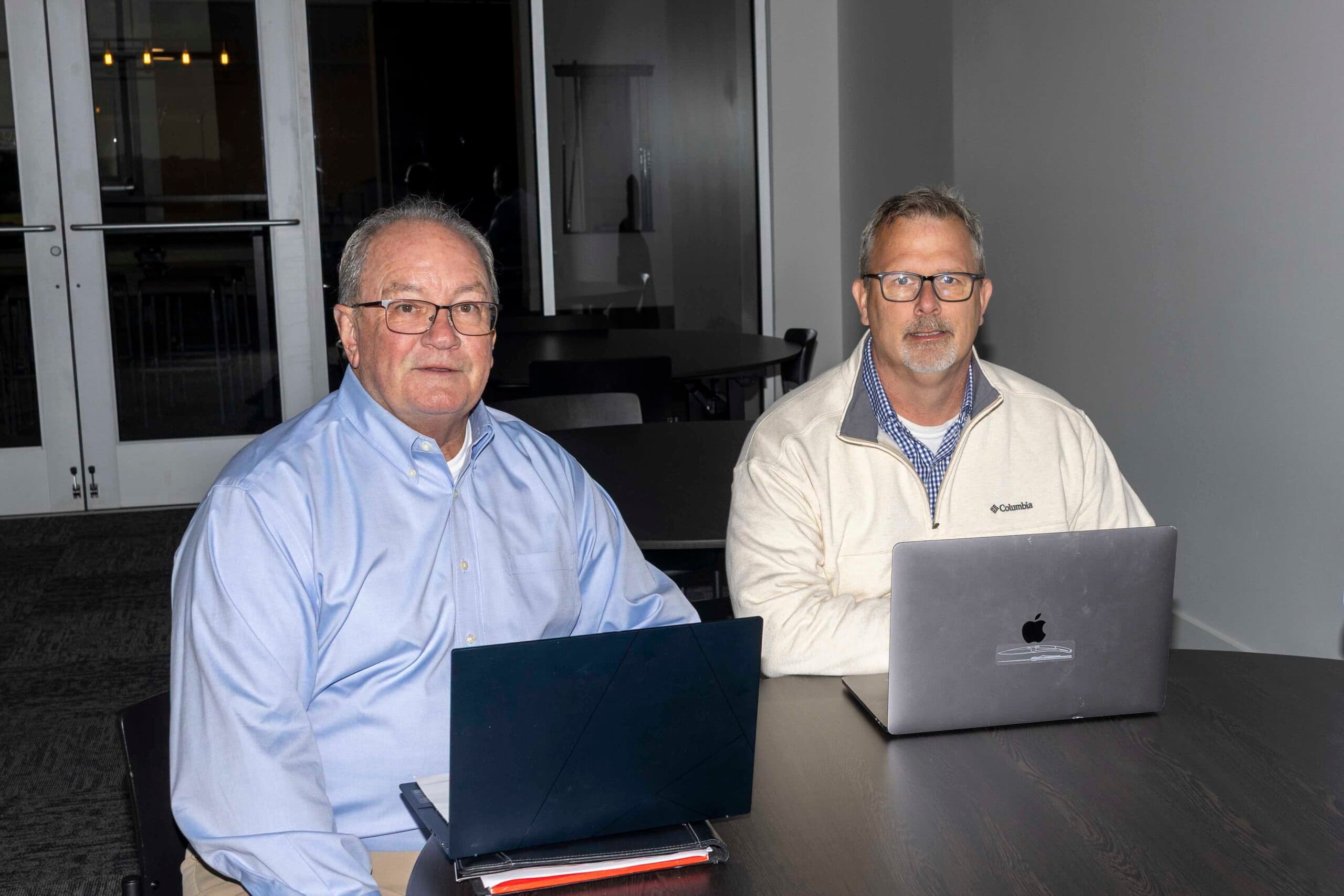 Two business brokers seated at a conference table with laptops in a modern office.